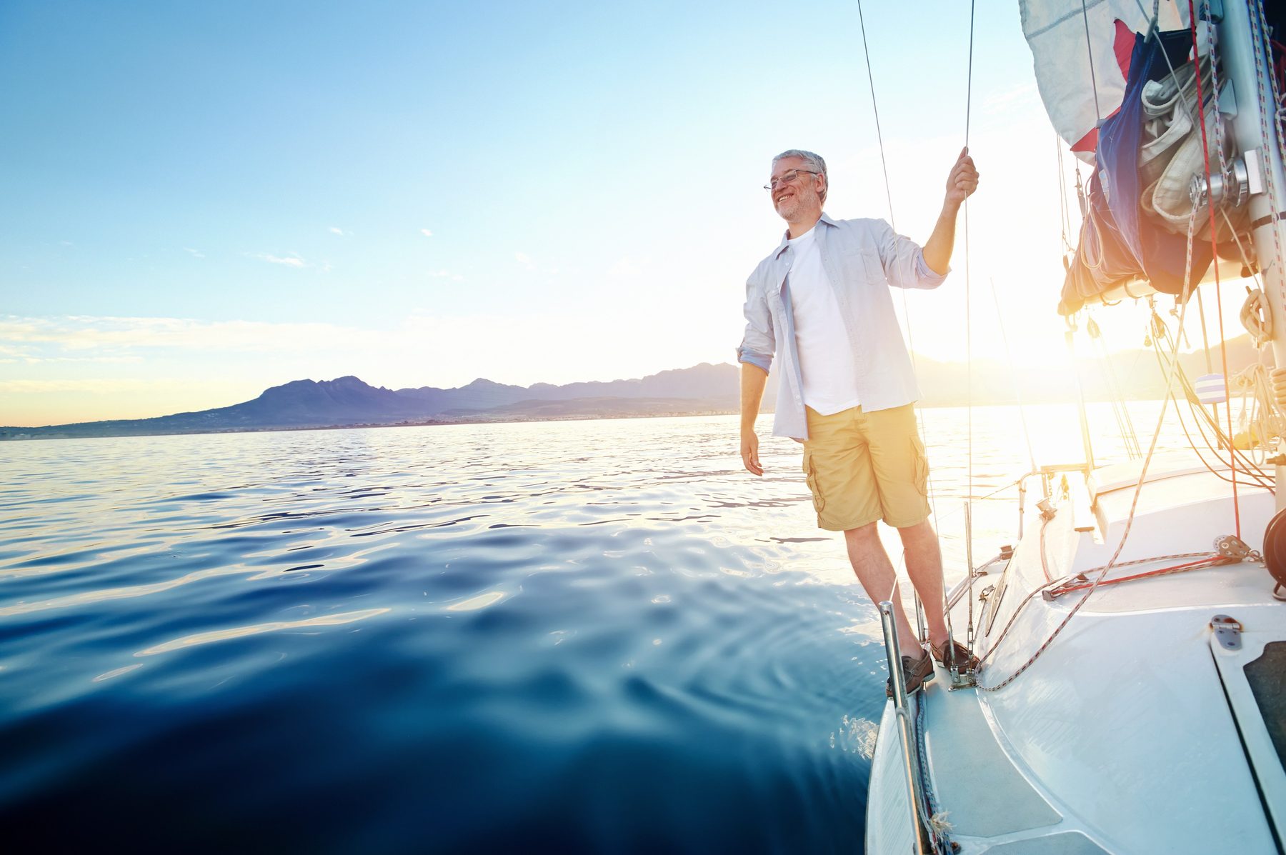 A man standing at the bow of a sailing boat, arm raised, looking out to sea at sunset