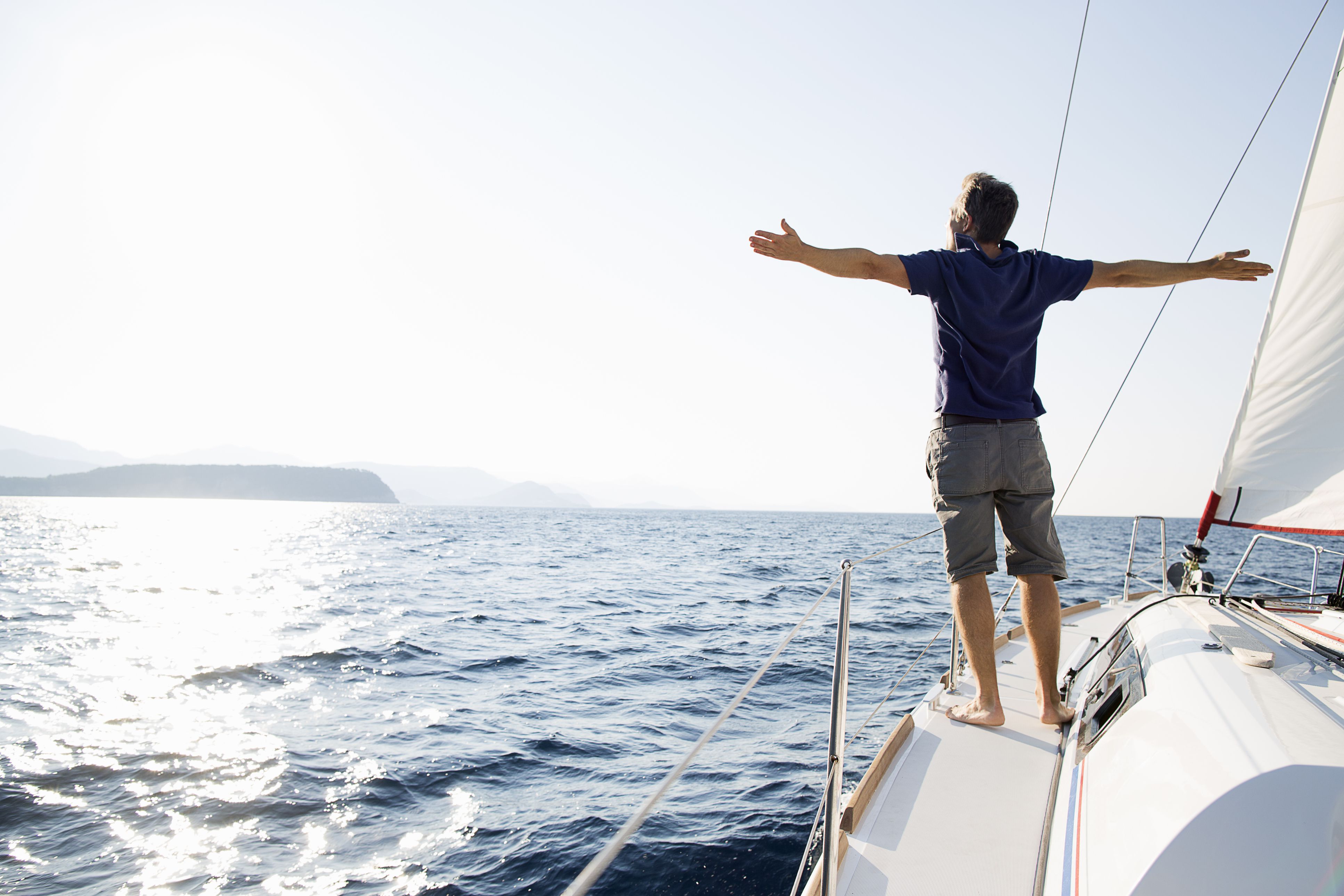 A man standing at the bow of a sailing boat, arms outstretched, looking out to sea
