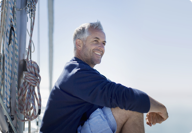 A man smiling and sitting on the deck of a sailing boat, surrounded by rope and rigging